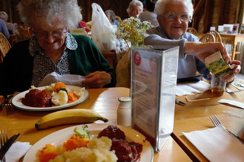 Vanhat iloiset silmälasipäiset rouvat istuvat syömässä pöydän ääressä salissa, jossa muitakin aterioimassa.